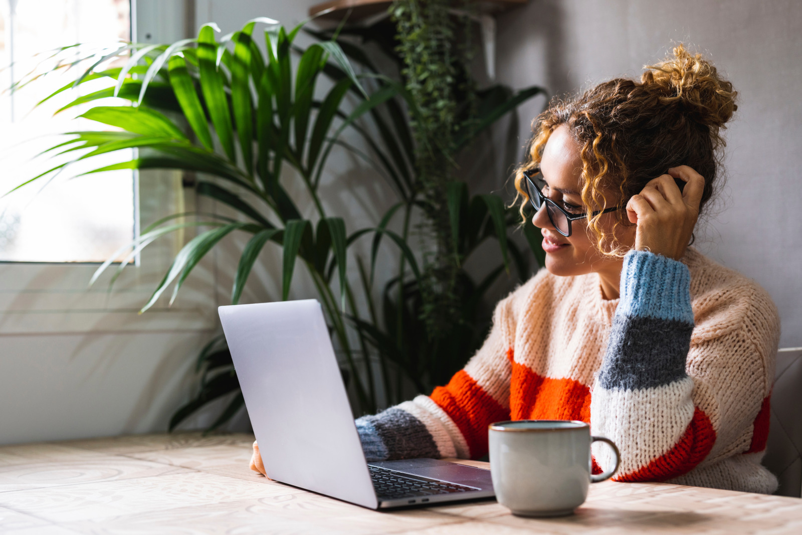 Femme adulte à la maison, surfant sur Internet avec son ordinateur portable posé sur une table, avec une grande plante en arrière-plan. Loisirs technologiques à la maison.